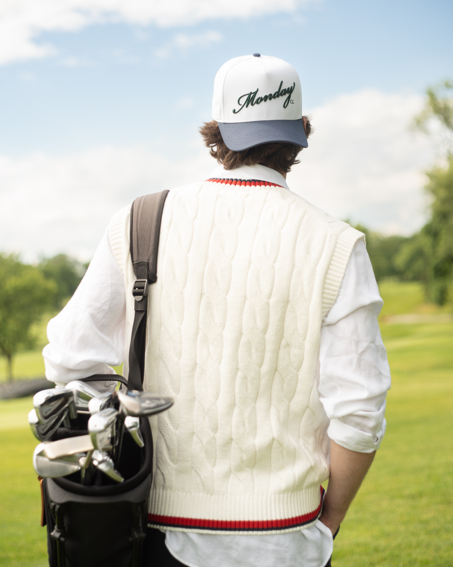Person on a golf course wearing a white sweater vest and cap, carrying golf clubs.