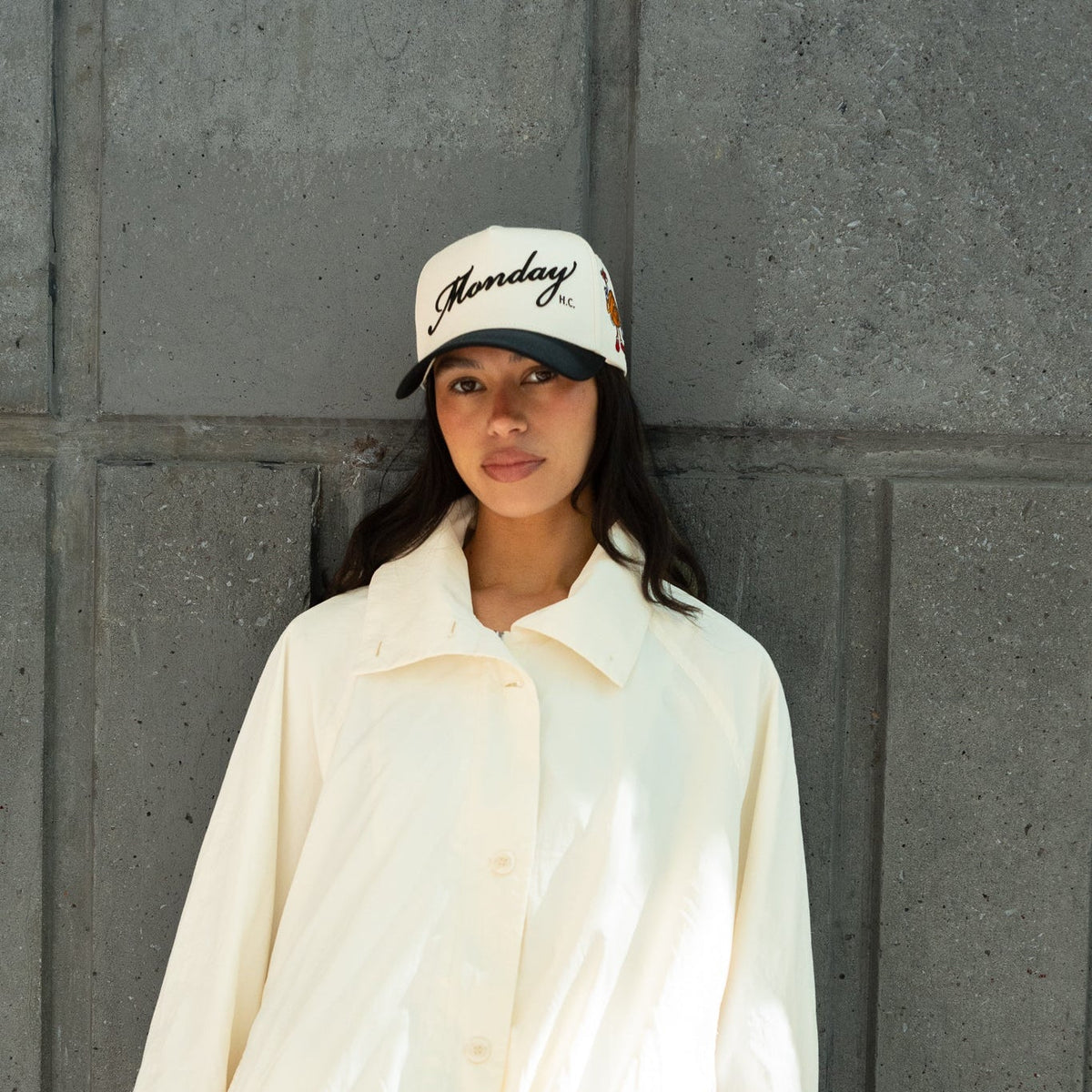 A woman wearing a cream-colored lightweight jacket and matching pants, paired with a white baseball cap that reads 'Monday', standing against a dark gray paneled wall.