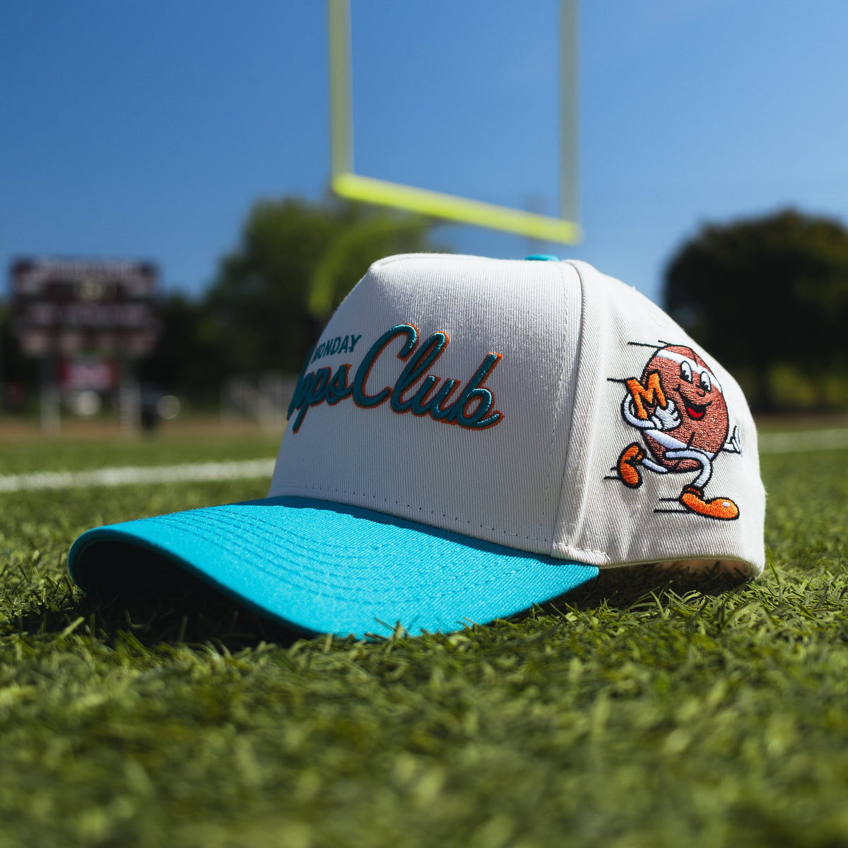A white and turquoise snapback cap with 'Texas A&M Aggie Club' embroidered on the front, resting on green grass with a football goalpost in the background.